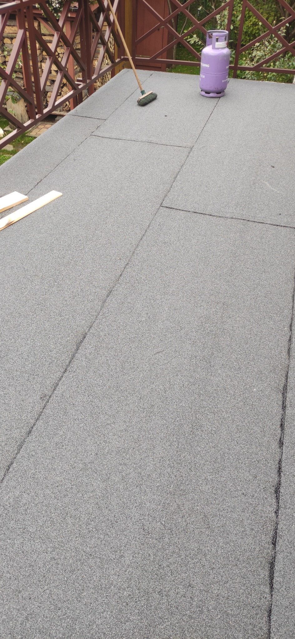 Nowe pokrycie dachu z papy bitumicznej w kolorze szarym, widoczna butla gazowa i szczotka dekarska na powierzchni, balustrada w tle.