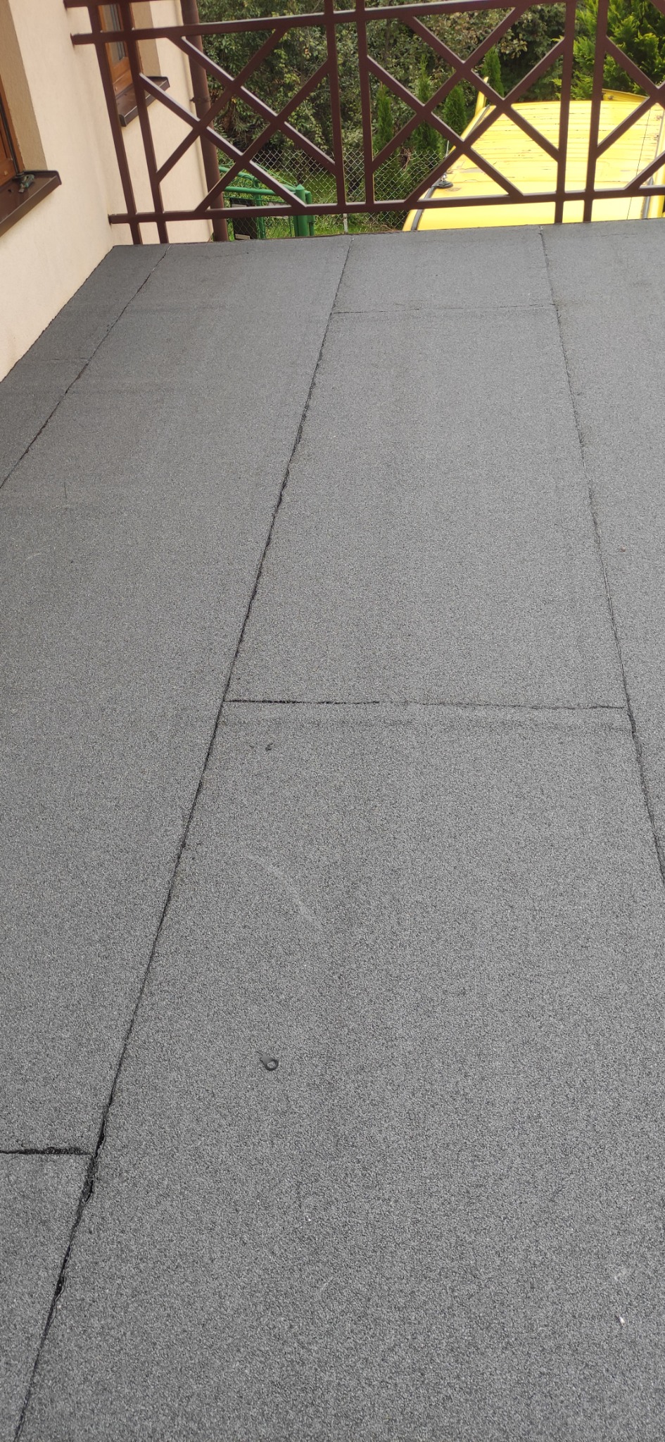 Szara, bitumiczna papa na dachu balkonu, widoczna z góry, z brązową balustradą w tle i fragmentem żółtego dachu budynku sąsiedniego.