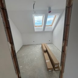 KOZ-BUD - Poddasze w trakcie remontu, widoczne dwa okna dachowe, ściany pomalowane na biało, na podłodze leżą kartony z materiałami budowlanymi, widoczna surowa ościeżnica drzwi.