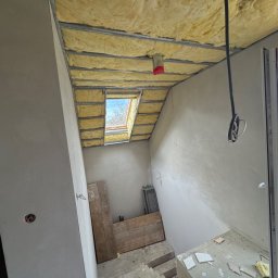 KOZ-BUD - Poddasze w trakcie ocieplania wełną mineralną, widoczne metalowe profile, okno dachowe i fragment balkonu z widokiem na krajobraz.