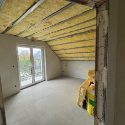 KOZ-BUD - Poddasze w trakcie ocieplania wełną mineralną, widoczne metalowe profile, okno balkonowe i częściowo wykończone ściany.