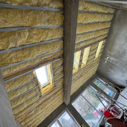 KOZ-BUD - Poddasze w trakcie ocieplania wełną mineralną, widoczne profile metalowe, okno dachowe oraz surowy beton na ścianach.