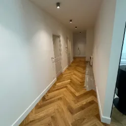 Korytarz z parkietem ułożonym w jodełkę, białe ściany z listwami przypodłogowymi, sufit z trzema lampami punktowymi. Częściowo widoczny fragment podłogi przed wykończeniem.