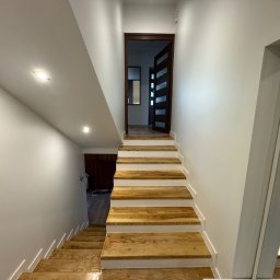 KOZ-BUD - Jasne schody z drewnianymi stopniami, prowadzące w dół, oświetlone punktowymi lampami sufitowymi, z widocznym fragmentem lustra po prawej stronie.