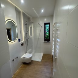 KOZ-BUD - Nowoczesna łazienka z kabiną prysznicową typu walk-in, podwieszaną toaletą i lustrem z podświetleniem LED. Uwagę zwraca okno z widokiem na zieleń.