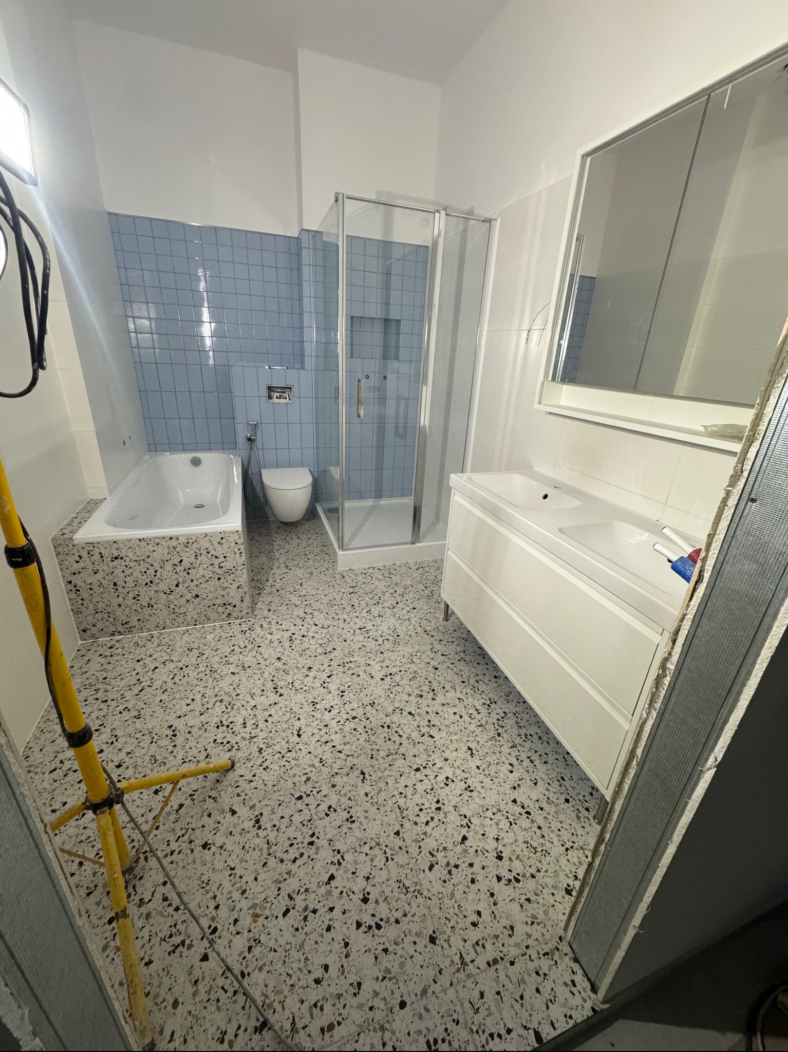 Jasna łazienka w trakcie wykańczania: wanna z obudową w terrazzo, kabina prysznicowa, umywalka z szafką, niebieskie płytki na ścianie, podłoga lastryko, widoczna lampa budowlana i profil metalowy.