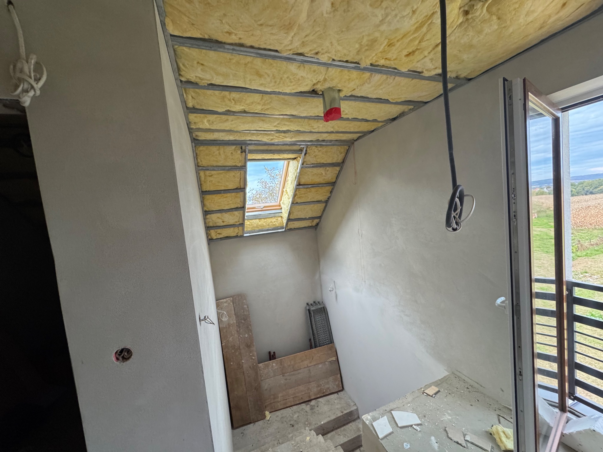 Poddasze w trakcie ocieplania wełną mineralną, widoczne metalowe profile, okno dachowe i fragment balkonu z widokiem na krajobraz.