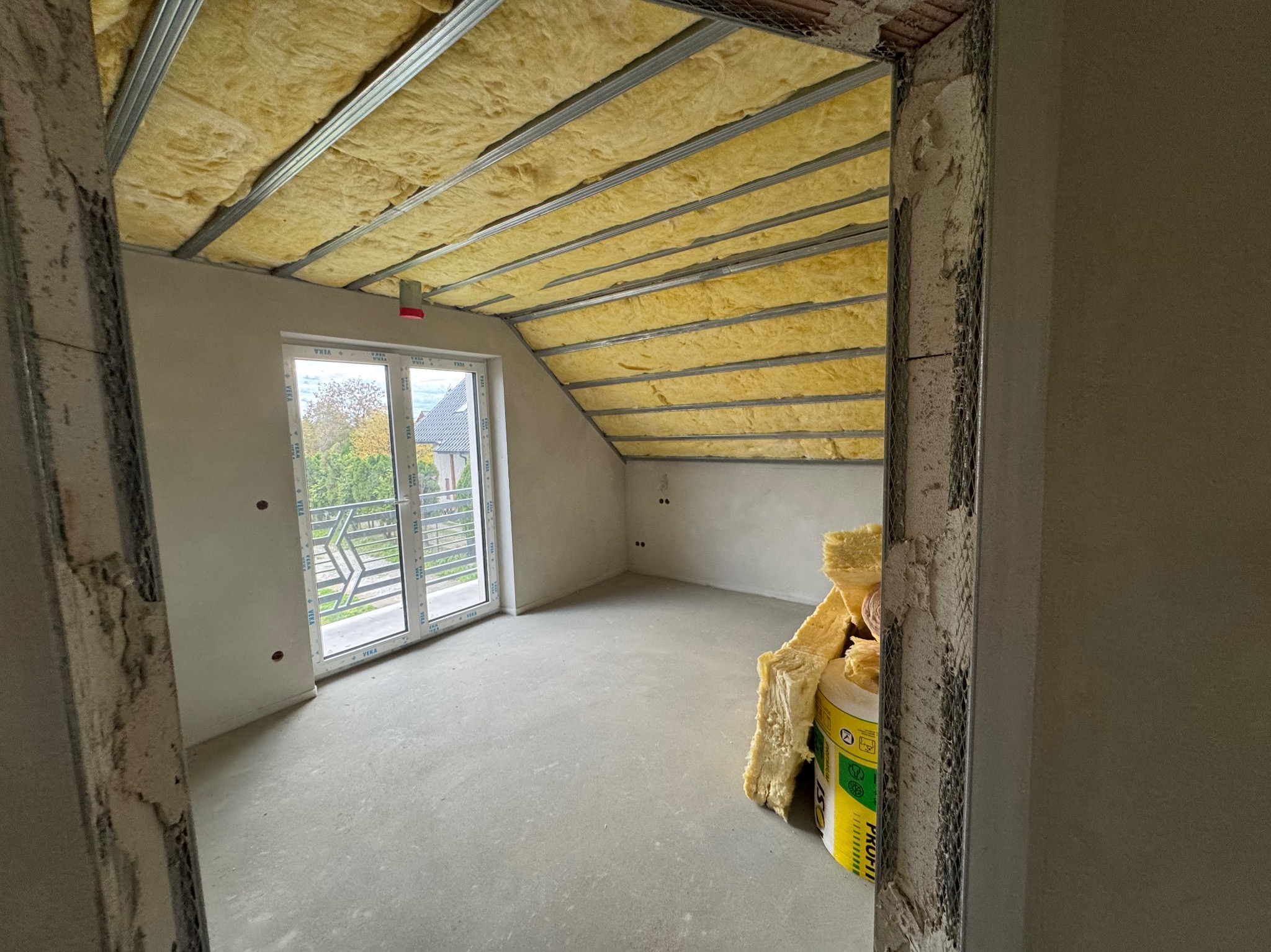 Poddasze w trakcie ocieplania wełną mineralną, widoczne metalowe profile, okno balkonowe i częściowo wykończone ściany.