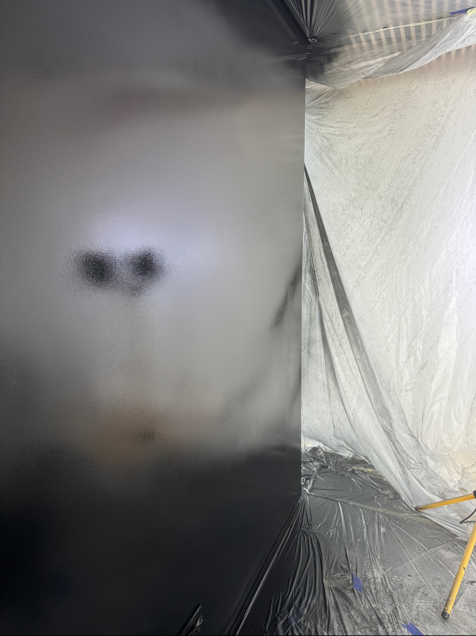 Świeżo pomalowana na czarno ściana, zabezpieczona folią malarską, z widocznym oświetleniem budowlanym.