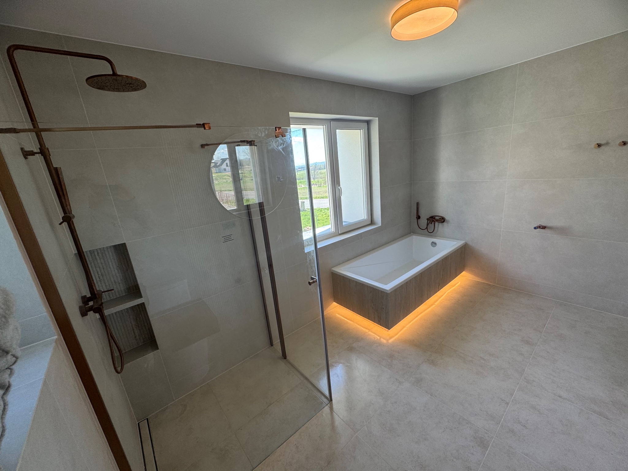 Nowoczesna łazienka z beżowymi płytkami, przeszklonym prysznicem i podświetlaną wanną przy oknie. Armatura miedziana. Minimalistyczny design.