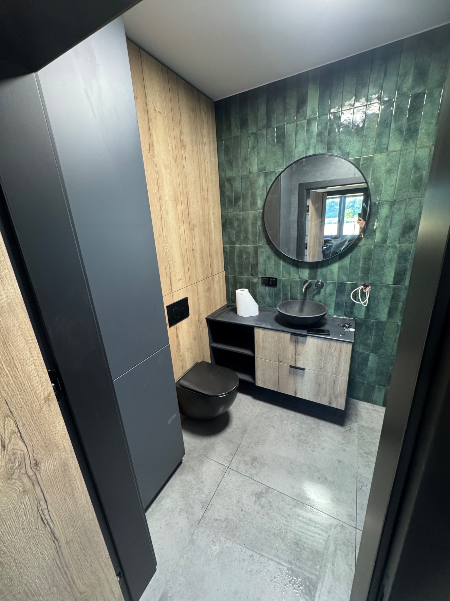 Nowoczesna łazienka z zielonymi płytkami, okrągłym lustrem, drewnianymi akcentami i szarą podłogą. Uwagę zwraca minimalistyczna umywalka i wisząca toaleta.