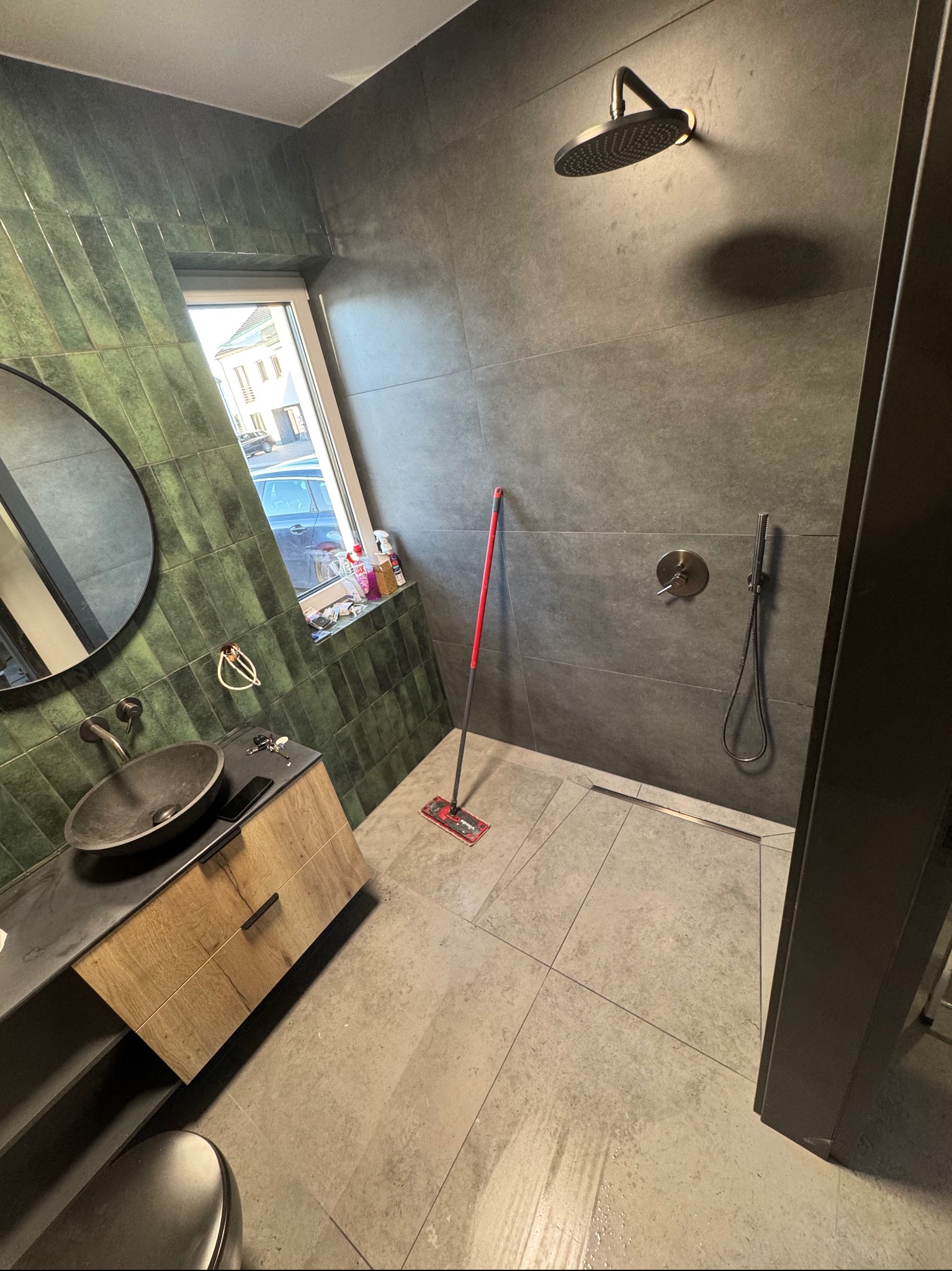 Nowoczesna łazienka z zielonymi i szarymi płytkami, deszczownicą, umywalką na szafce i oknem. Mop stoi na podłodze.