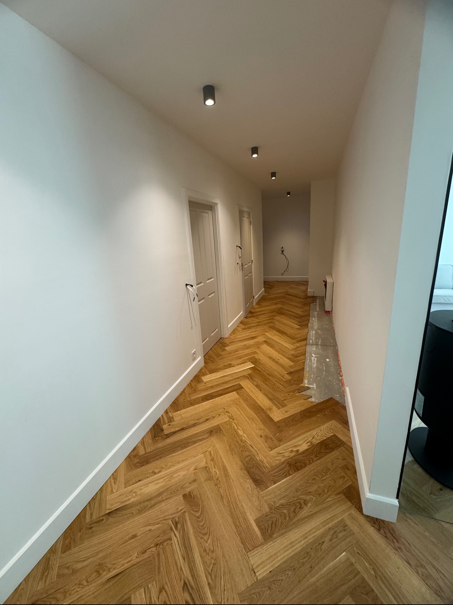 Korytarz z parkietem ułożonym w jodełkę, białe ściany z listwami przypodłogowymi, sufit z trzema lampami punktowymi. Częściowo widoczny fragment podłogi przed wykończeniem.