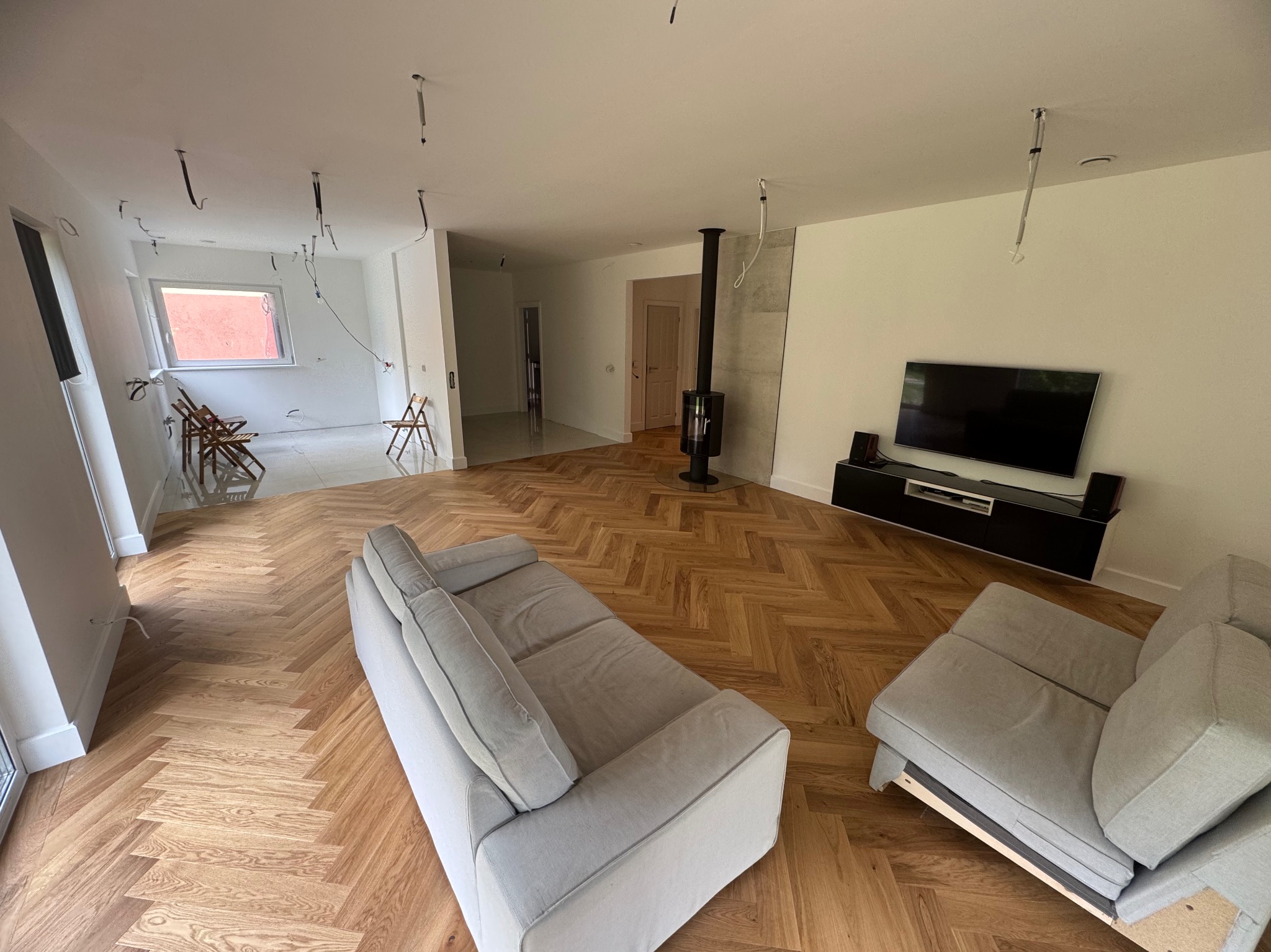 Salon z parkietem w jodełkę, szarą sofą i kominkiem. Część pomieszczenia w trakcie wykańczania, widoczne kable i surowe ściany. Minimalistyczny design z elementami w stylu industrialnym.