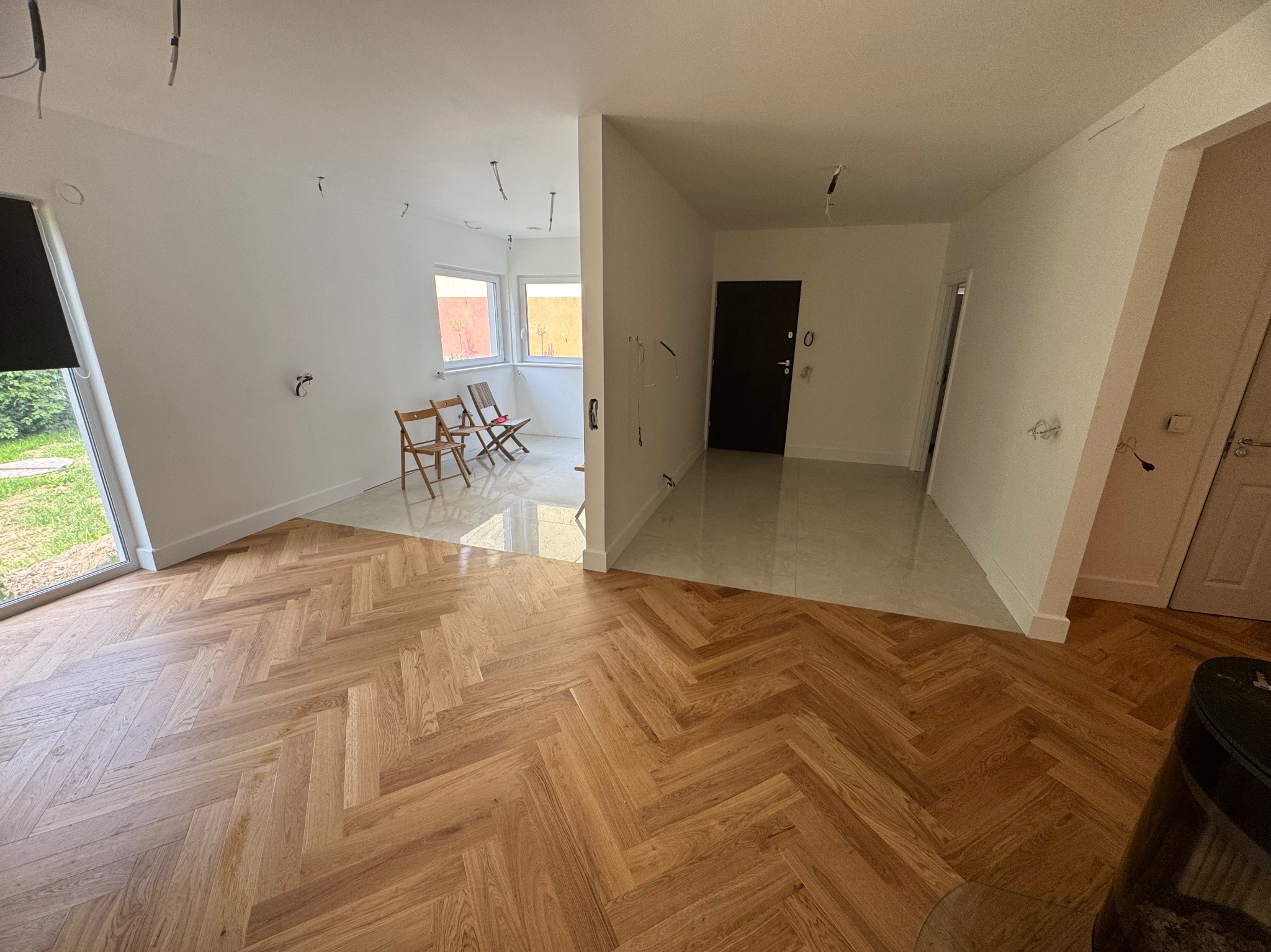 Jasny parkiet ułożony w jodełkę, przechodzący w jasne płytki. W tle białe ściany, okna i drzwi. Widoczne kable wystające z sufitu. Wnętrze w trakcie wykańczania.