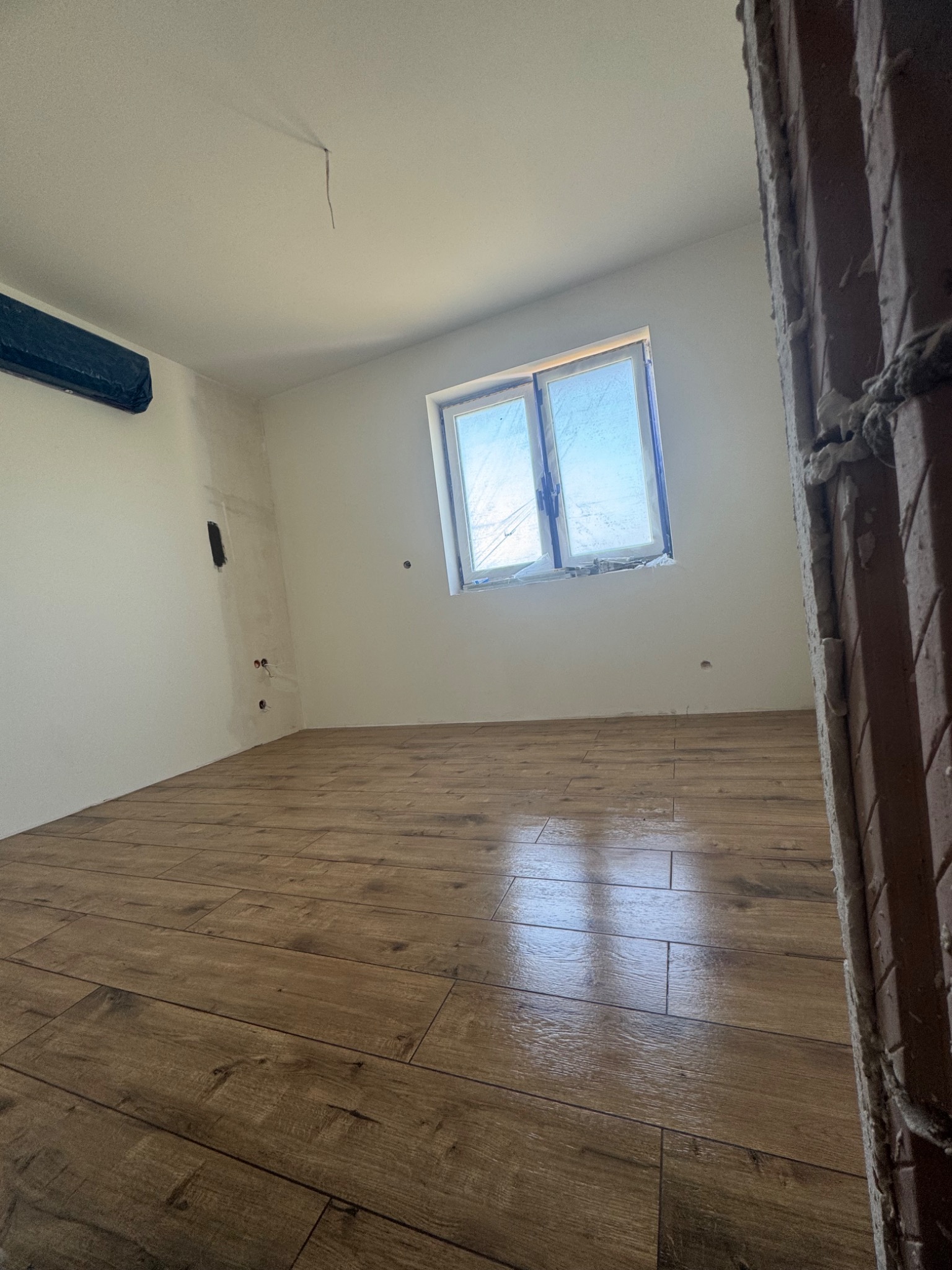 Pokój z ułożoną podłogą z paneli imitujących drewno. Ściany pomalowane na biało, okno zabezpieczone folią. Fragment surowego muru w prawym dolnym rogu.