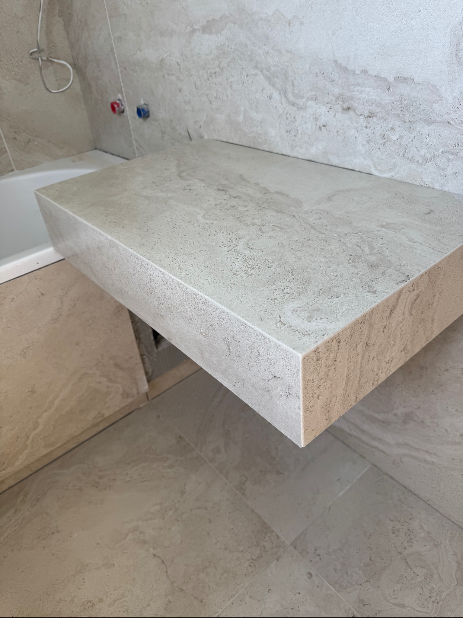 Wykończona łazienka: beżowa, kamienna umywalka wisząca na tle płytek imitujących kamień, widoczne baterie i fragment wanny. Minimalistyczny design.