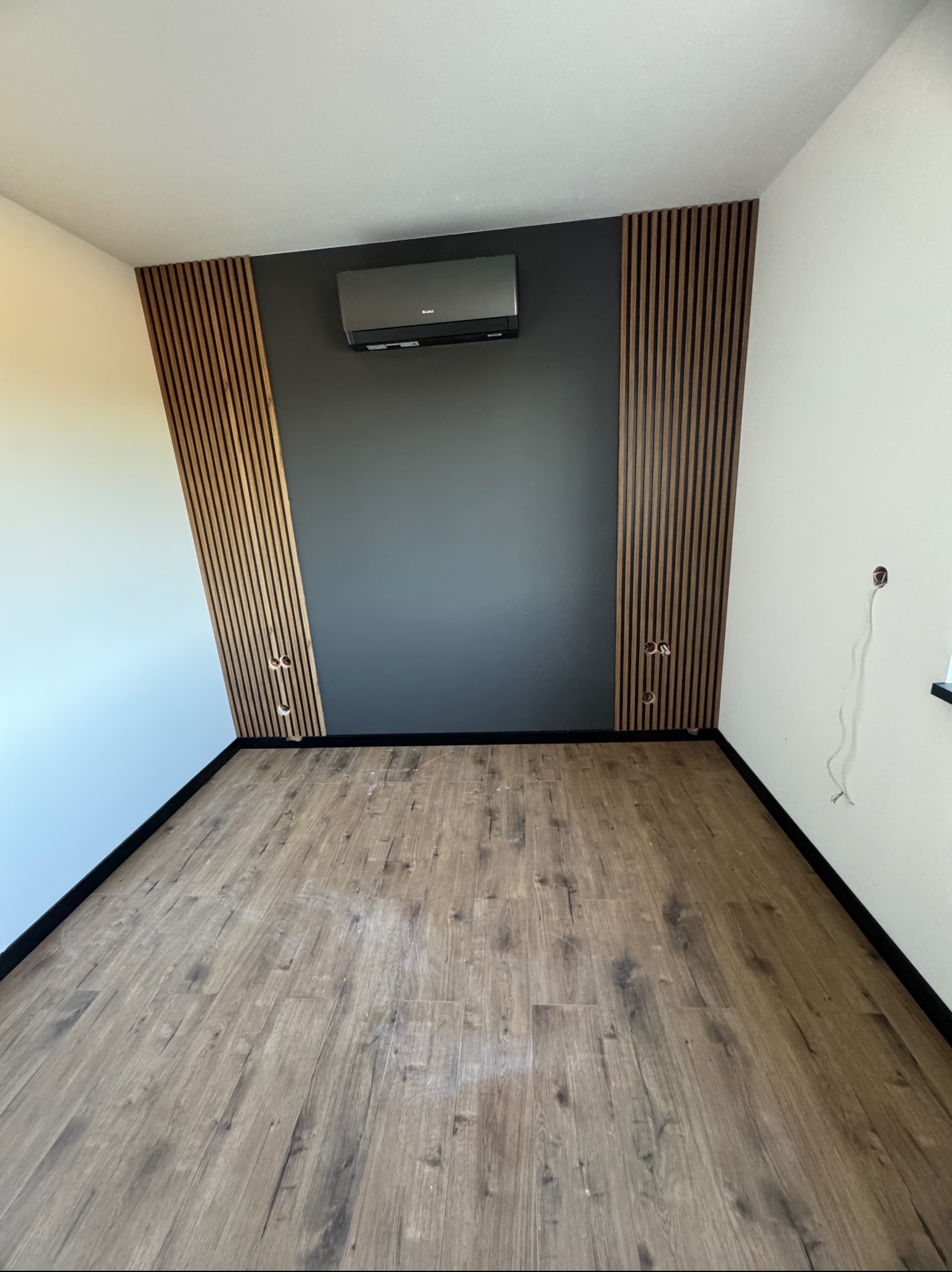 Nowoczesne wnętrze z drewnianymi lamelami na ścianie, szarą farbą i klimatyzacją. Jasna podłoga z paneli, widoczne gniazdka elektryczne.
