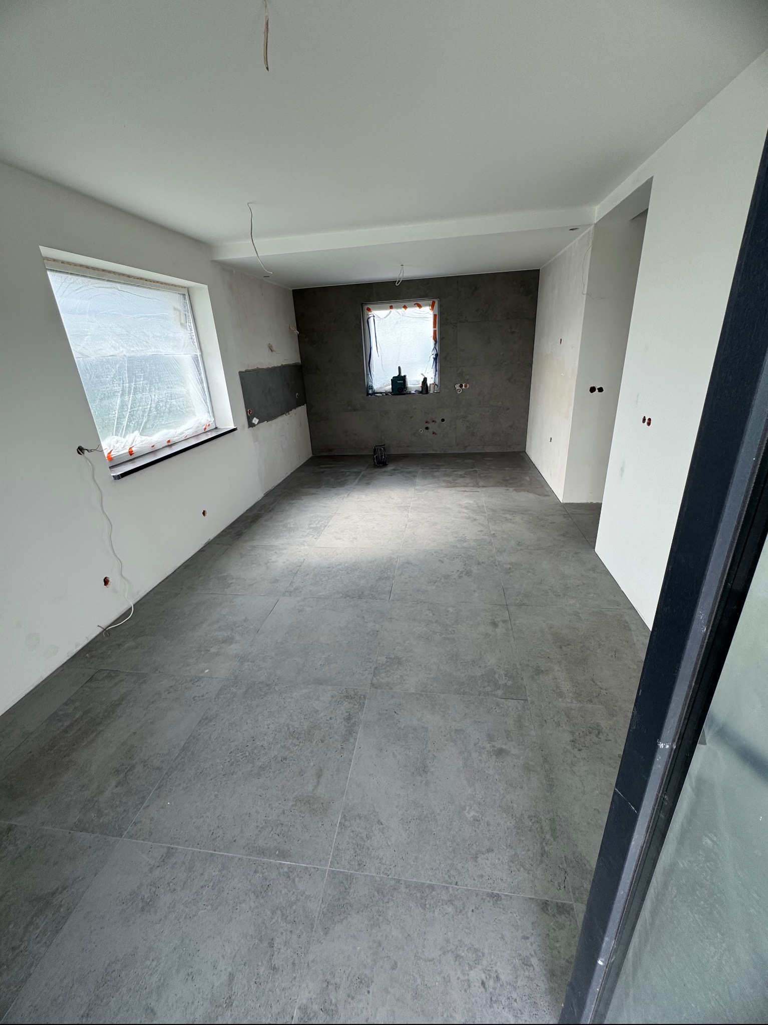 Wnętrze w trakcie prac: szare płytki na podłodze, częściowo wykończone ściany, okna zabezpieczone folią. Widoczne punkty elektryczne i surowe wykończenie.