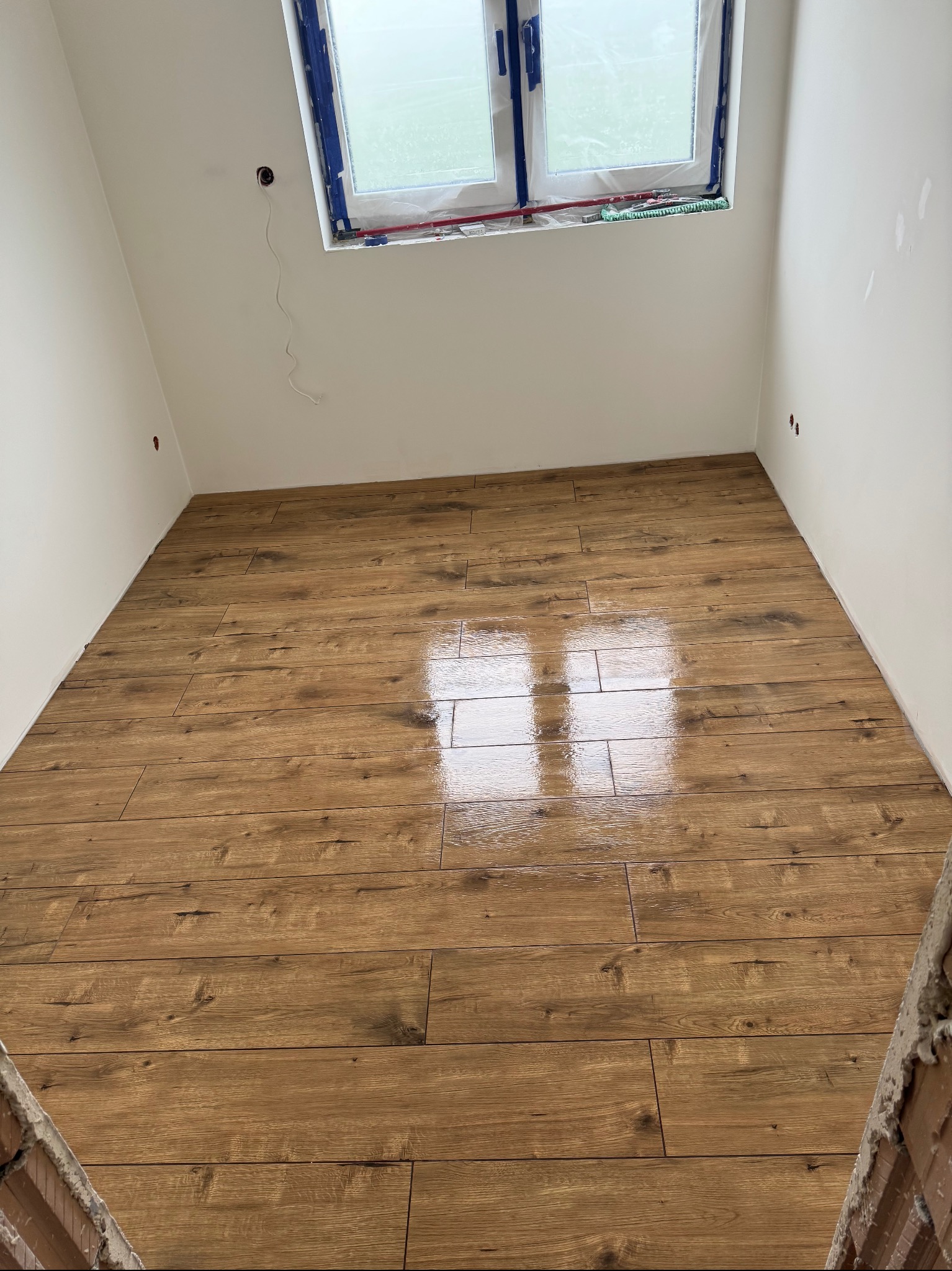 Jasne wnętrze z ułożoną podłogą z płytek imitujących drewno. Widoczne białe ściany i okno z niebieską ramą. Podłoga błyszcząca, z odbiciem światła.