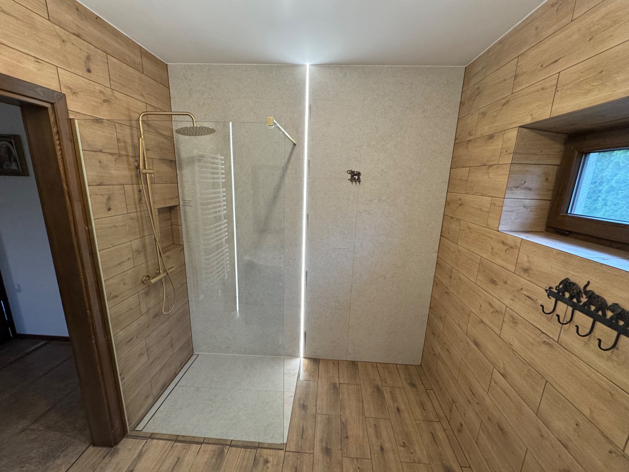 Nowoczesna łazienka z kabiną prysznicową typu walk-in, wykończona płytkami imitującymi drewno i subtelnym oświetleniem LED. Widoczne detale: złota armatura i dekoracyjny wieszak.