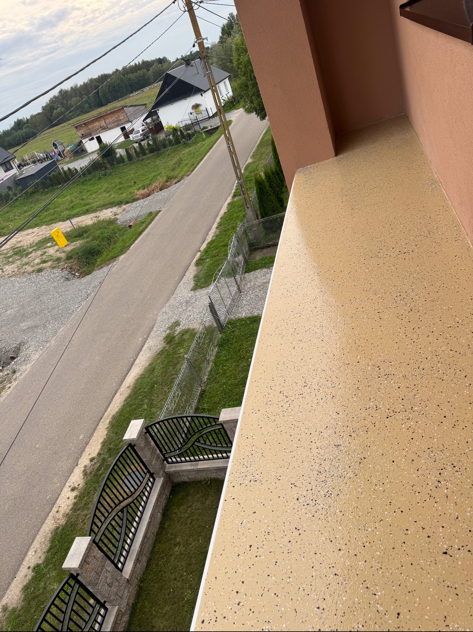 Balkon z beżową, żywiczną posadzką z ciemnymi drobinkami, widok z góry na ogrodzenie i ulicę. Nowoczesne wykończenie balkonu.