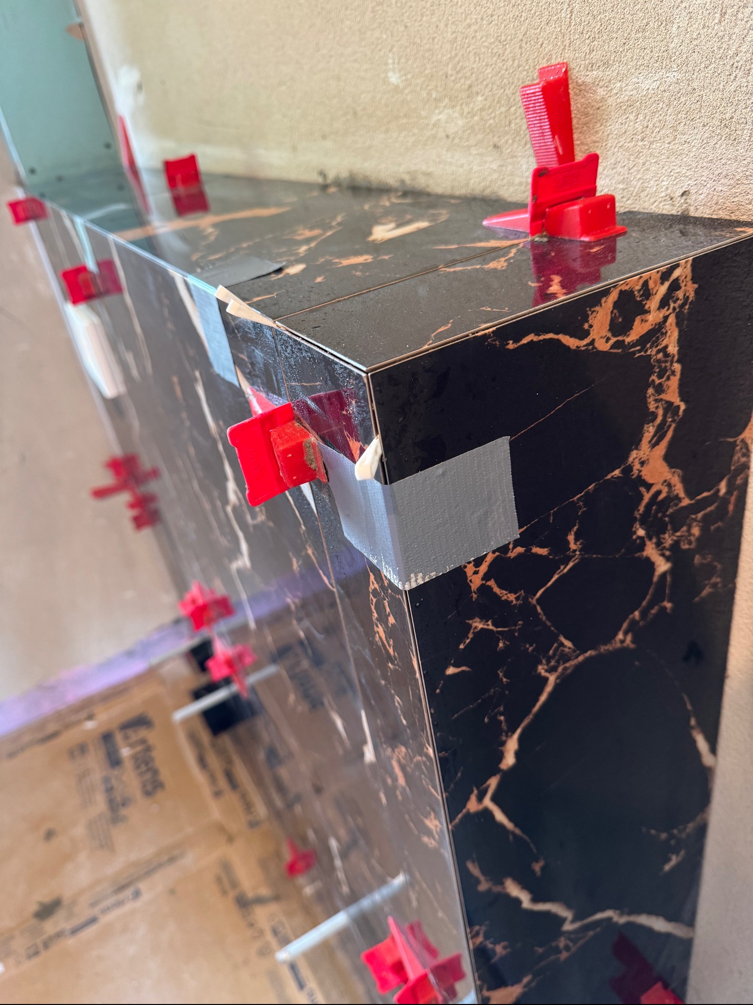 Montaż ciemnych płytek imitujących marmur z użyciem poziomujących klipsów i taśmy. Widoczny fragment pomieszczenia w trakcie prac wykończeniowych.