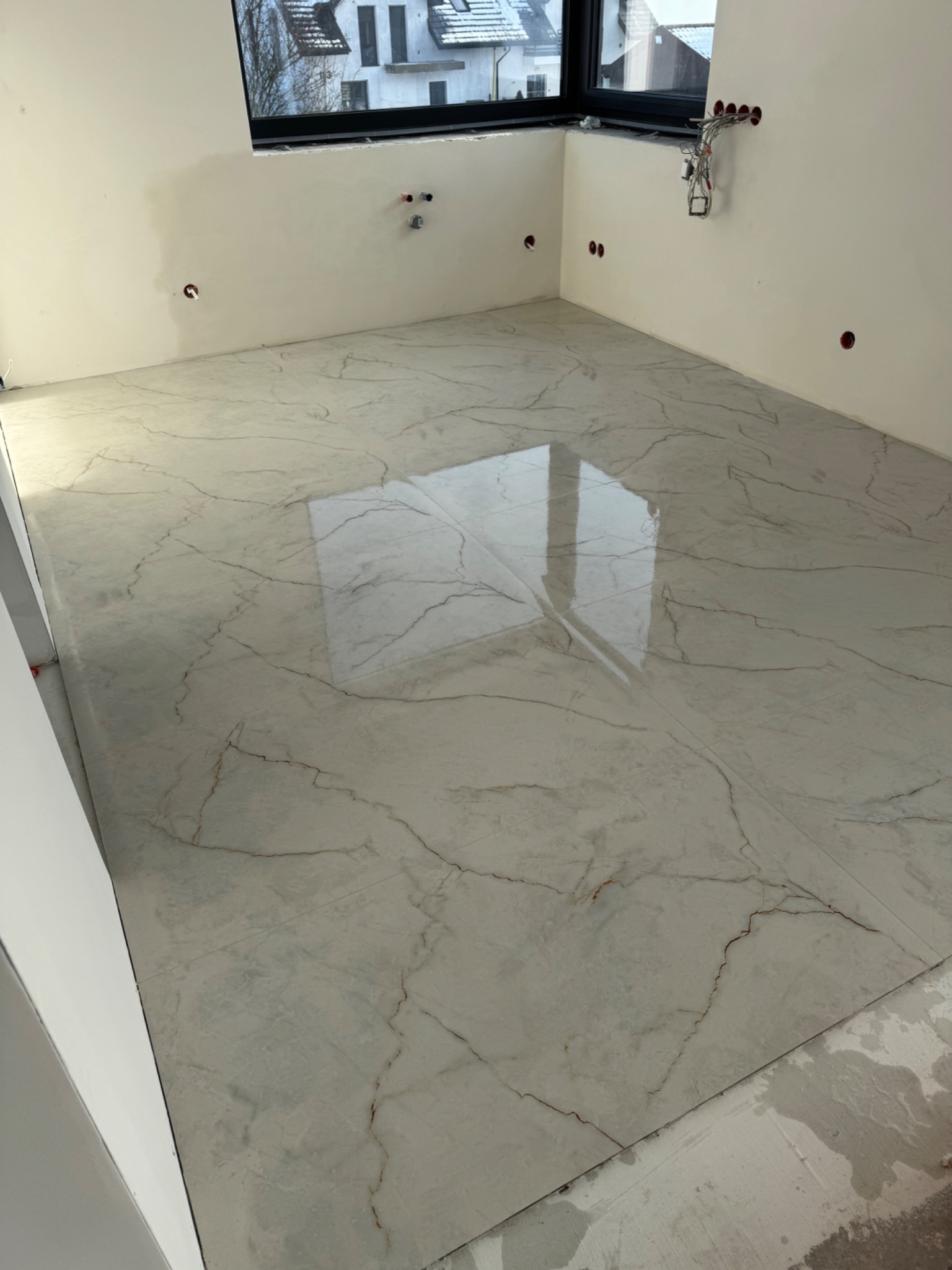 Błyszcząca, kremowa podłoga z marmurkowym wzorem w nowym pomieszczeniu. Widoczne przygotowane punkty instalacji na ścianach i okno z widokiem na zabudowę.