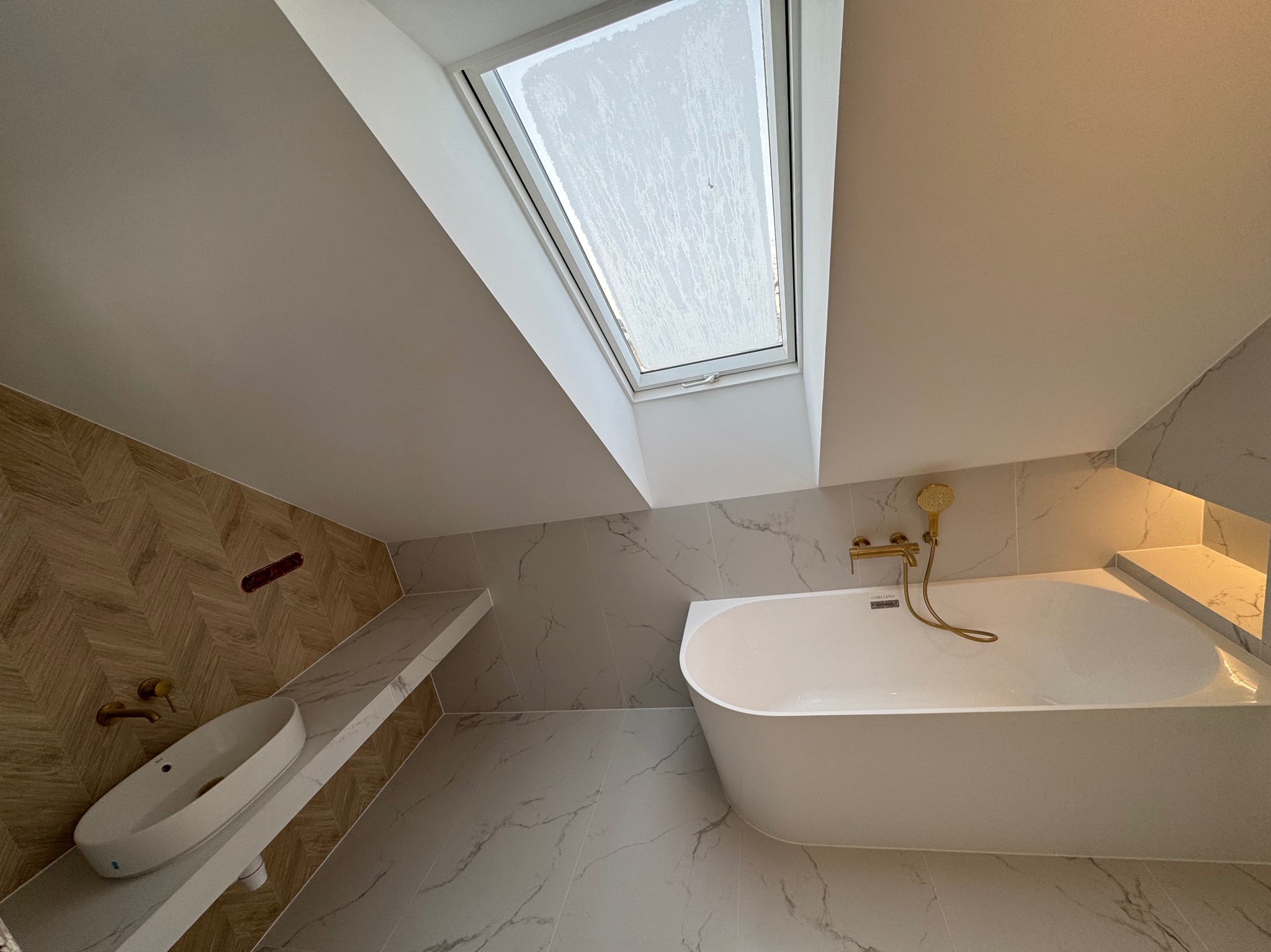 Nowoczesna łazienka na poddaszu z oknem dachowym, białą wanną i umywalką na blacie z marmurowym wzorem. Złota armatura i płytki imitujące drewno dodają elegancji.