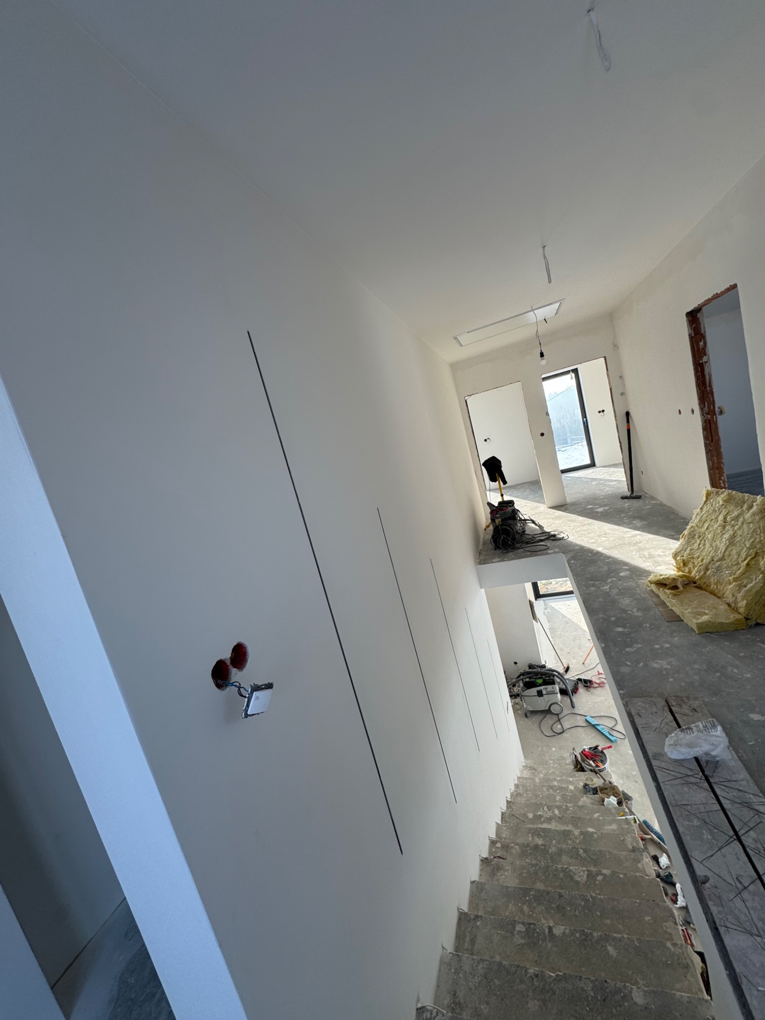 Wnętrze w trakcie prac wykończeniowych: białe ściany z widocznymi puszkami elektrycznymi, surowe schody i narzędzia budowlane. Widok z góry na korytarz.