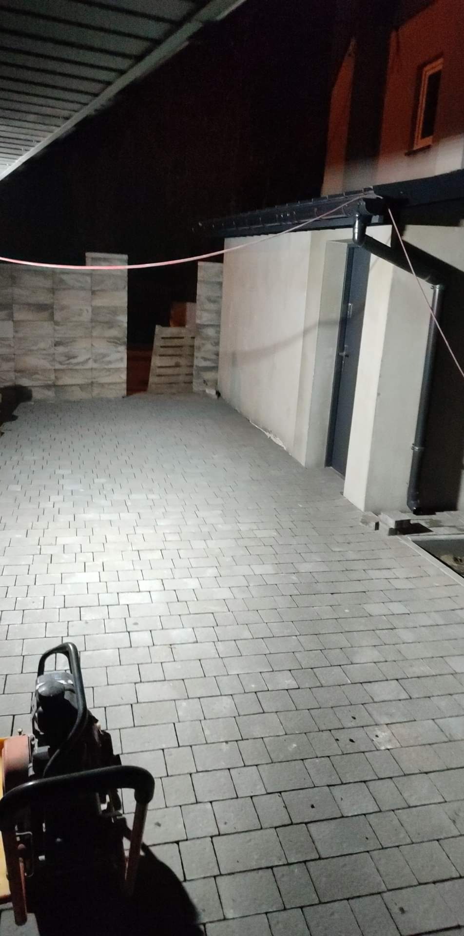 Ułożona szara kostka brukowa na posesji, widoczna ubijarka wibracyjna i paleta z kostką w tle, oświetlenie nocne.