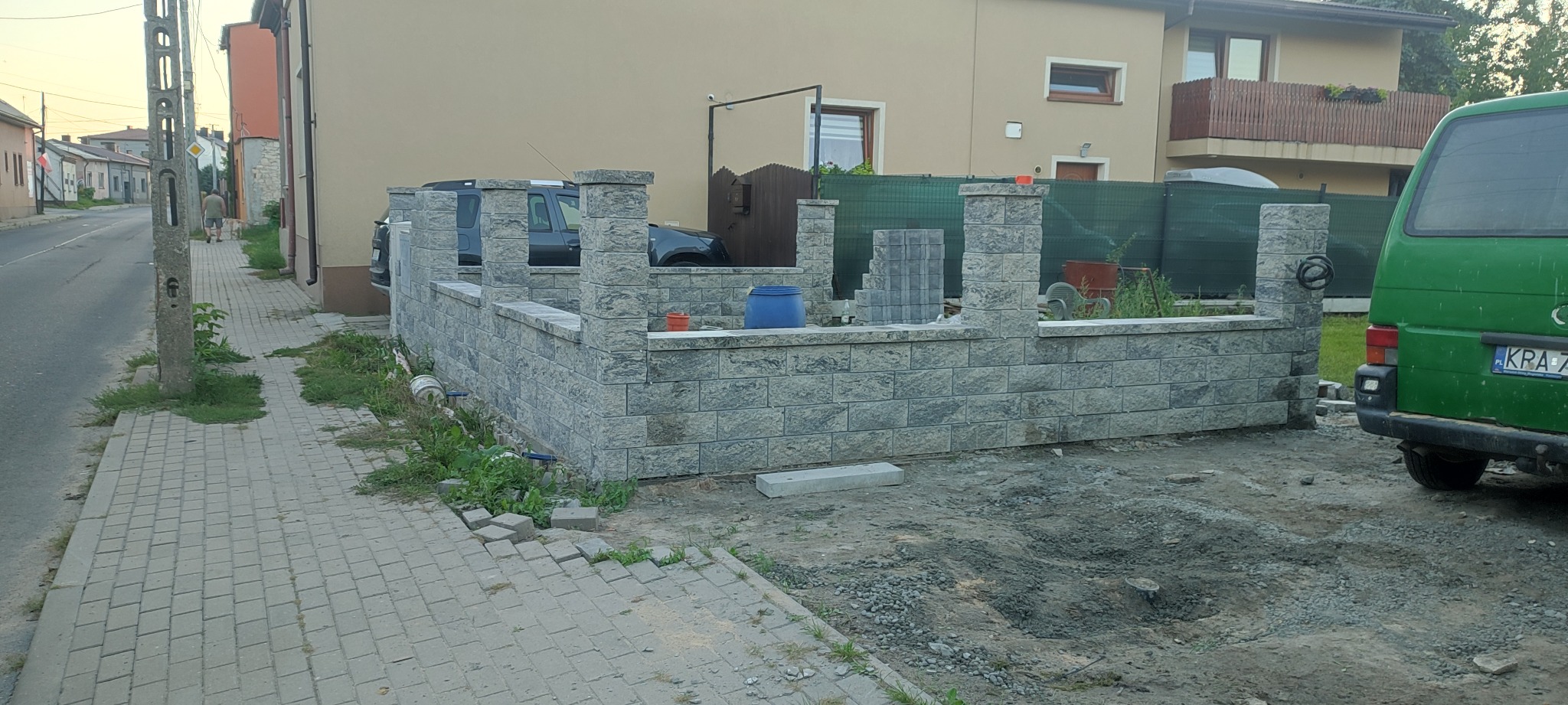 Wznoszony murek z szarej kostki łupanej, z widocznymi słupkami, na posesji przed domem, z zielonym busem w tle i częściowo przygotowanym podłożem.