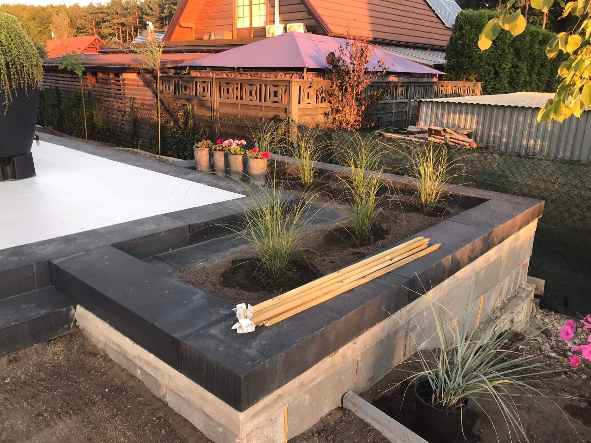 Kaskadowa rabata z ciemnej kostki, w trakcie budowy, z widocznymi elementami konstrukcyjnymi i przygotowanymi roślinami do nasadzenia, drewniane paliki na krawędzi.