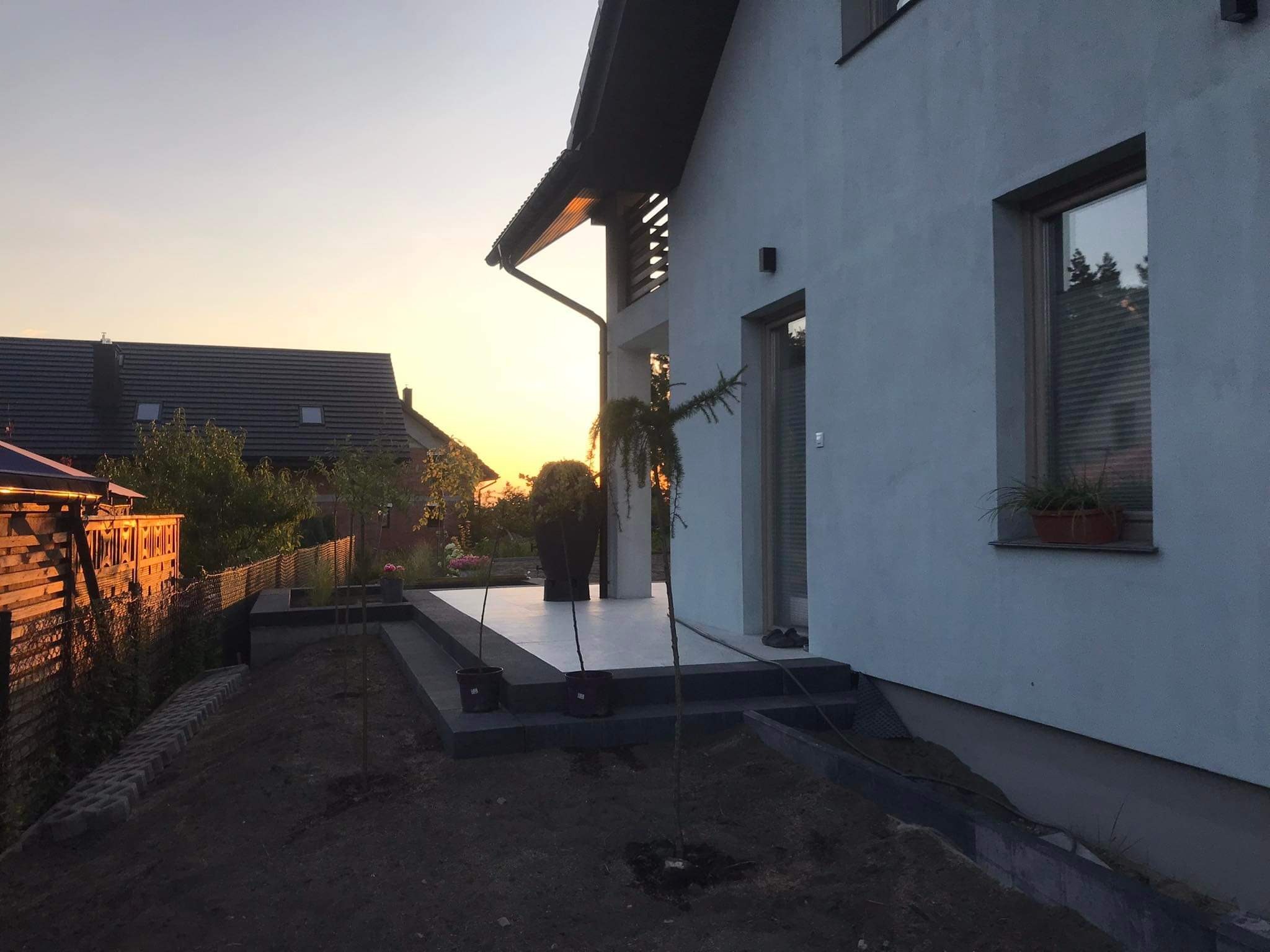 Nowoczesny taras z jasnych płyt, obrzeża z ciemnej kostki, młode drzewka posadzone w ziemi na tle domu o minimalistycznej architekturze, widok o zmierzchu.