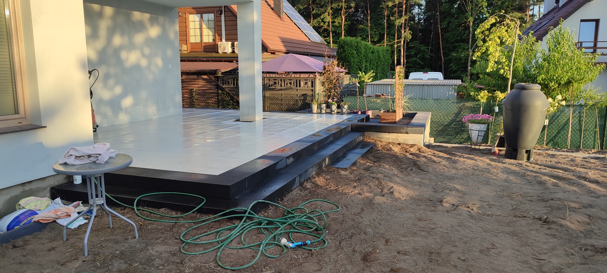 Biała kostka brukowa na tarasie domu jednorodzinnego z czarnymi obrzeżami i schodami, widoczny wąż ogrodowy, stół z rzeczami i duża donica.