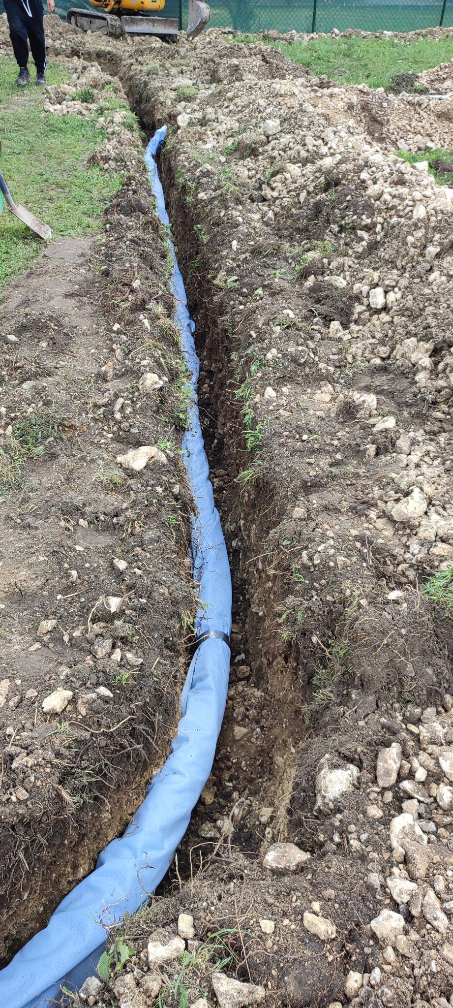 Wykop w ziemi z niebieską rurą zabezpieczoną taśmą, widoczny fragment koparki i pracownik z łopatą.