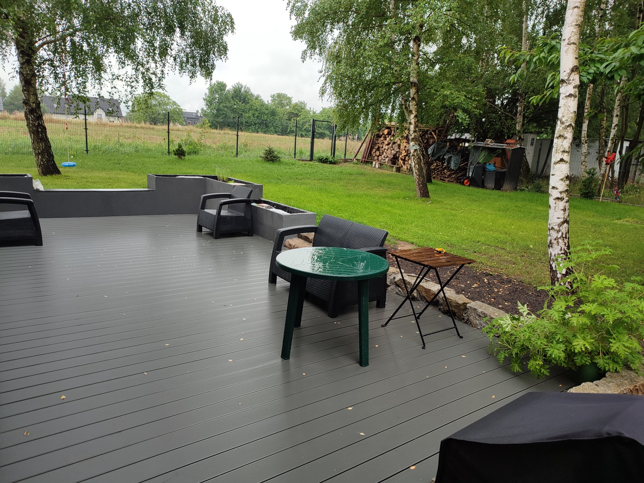 Szary taras kompozytowy z meblami ogrodowymi (krzesła rattanowe, zielony stolik) na tle trawnika z brzozami i stosem drewna.