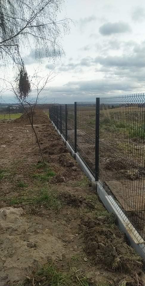 Długi, metalowy płot panelowy z betonową podmurówką, widoczny na tle zaoranego pola i pochmurnego nieba. Płot biegnie wzdłuż krawędzi działki, a obok niego leży świeżo usypana ziemia.