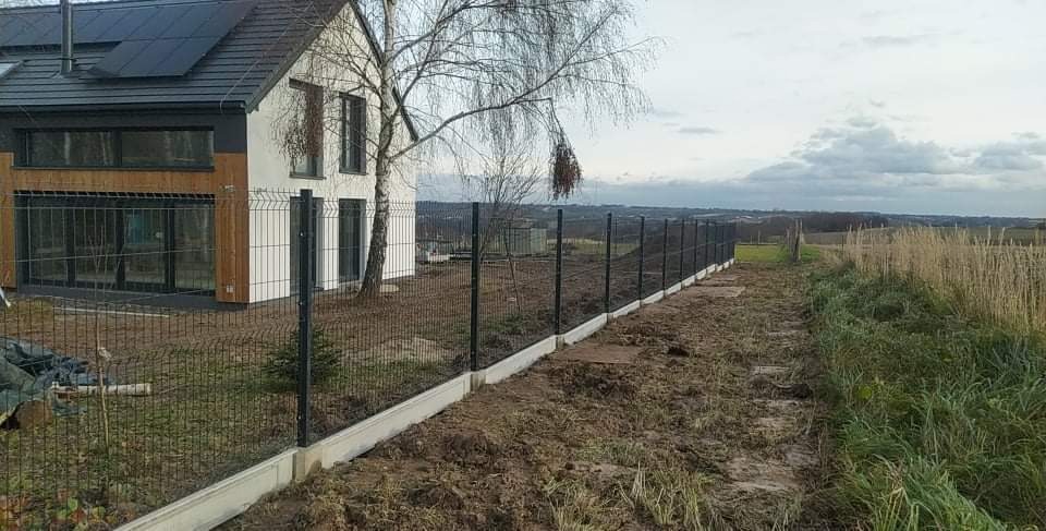 Nowoczesny dom z panelami słonecznymi w Wielkiej Wsi, otoczony ogrodzeniem panelowym z podmurówką betonową, widok na pole i zachmurzone niebo.