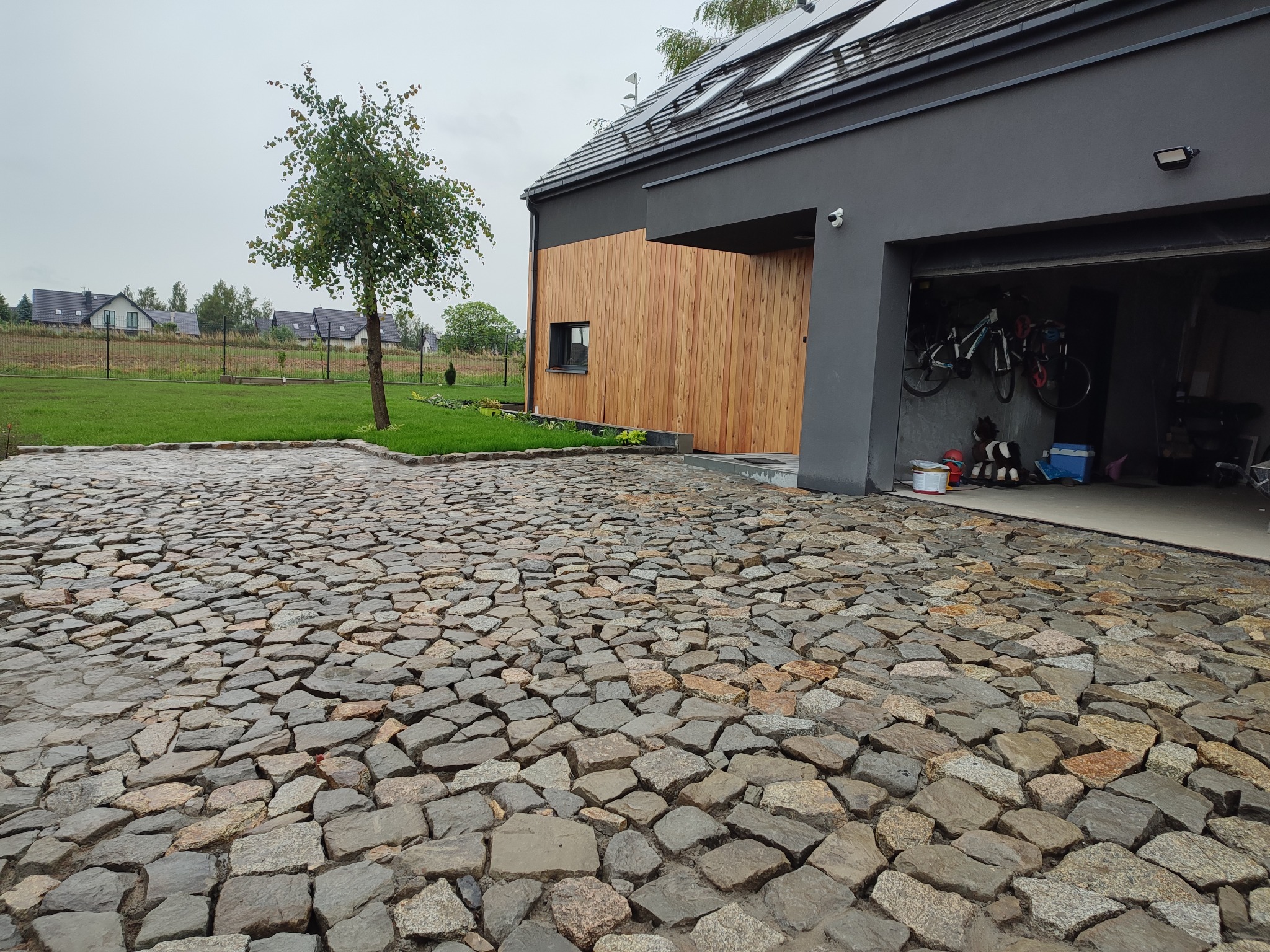 Ułożona kostka granitowa na podjeździe nowoczesnego domu z drewnianą elewacją, widok na otwarty garaż z rowerami i narzędziami w tle.