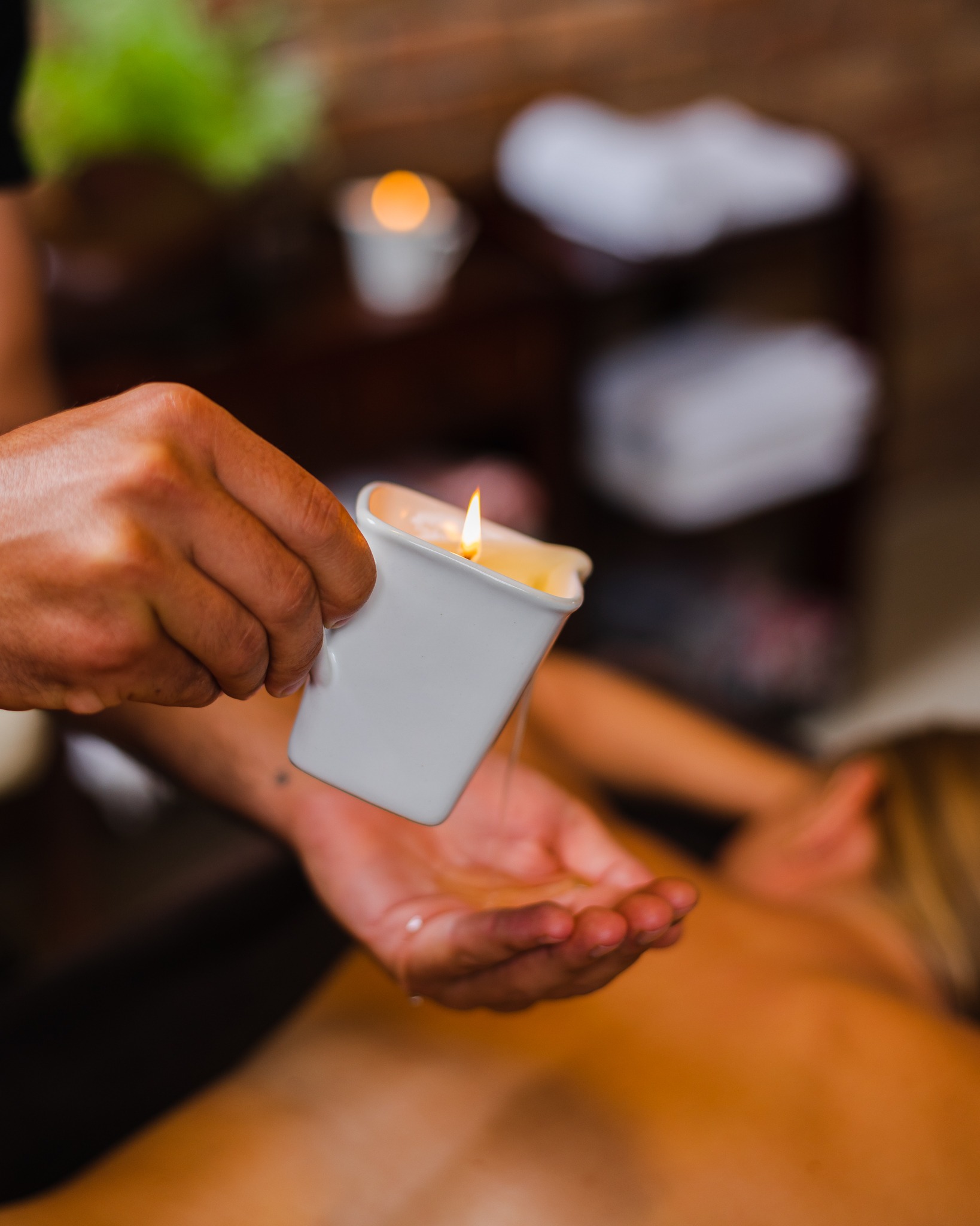 Olej do masażu spływa z białego naczynia z płonącą świeczką do dłoni masażysty, przygotowującego się do zabiegu na plecach klienta.