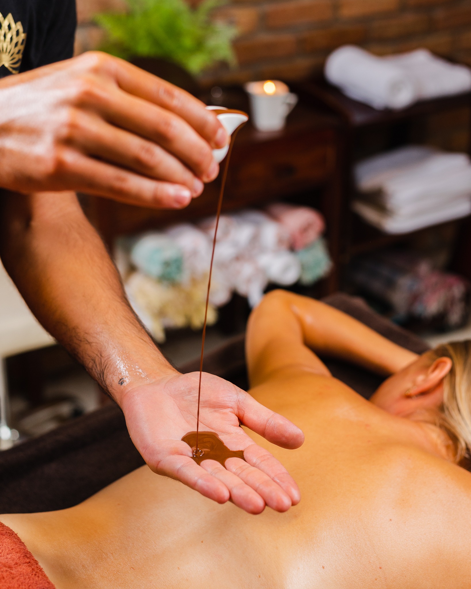 Masażysta w czarnej koszulce z złotym logo wlewa ciepłą czekoladę z białego naczynia na dłoń, przygotowując do masażu pleców kobiety leżącej na brązowym stole w gabinecie SPA, w tle świeca i ręczniki.