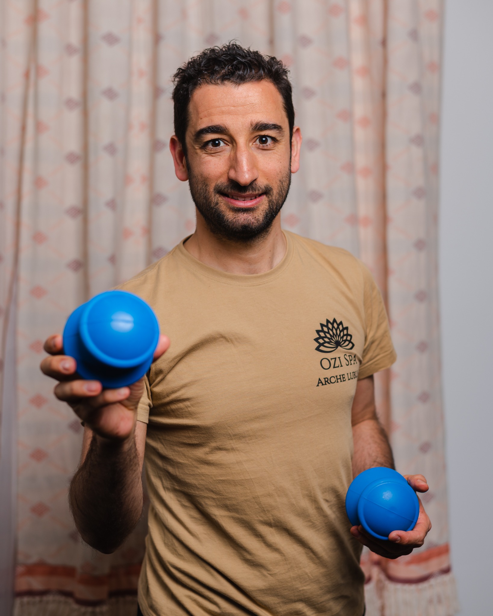 Uśmiechnięty mężczyzna w beżowej koszulce z logo, prezentujący niebieskie bańki do masażu próżniowego na tle wzorzystej zasłony.