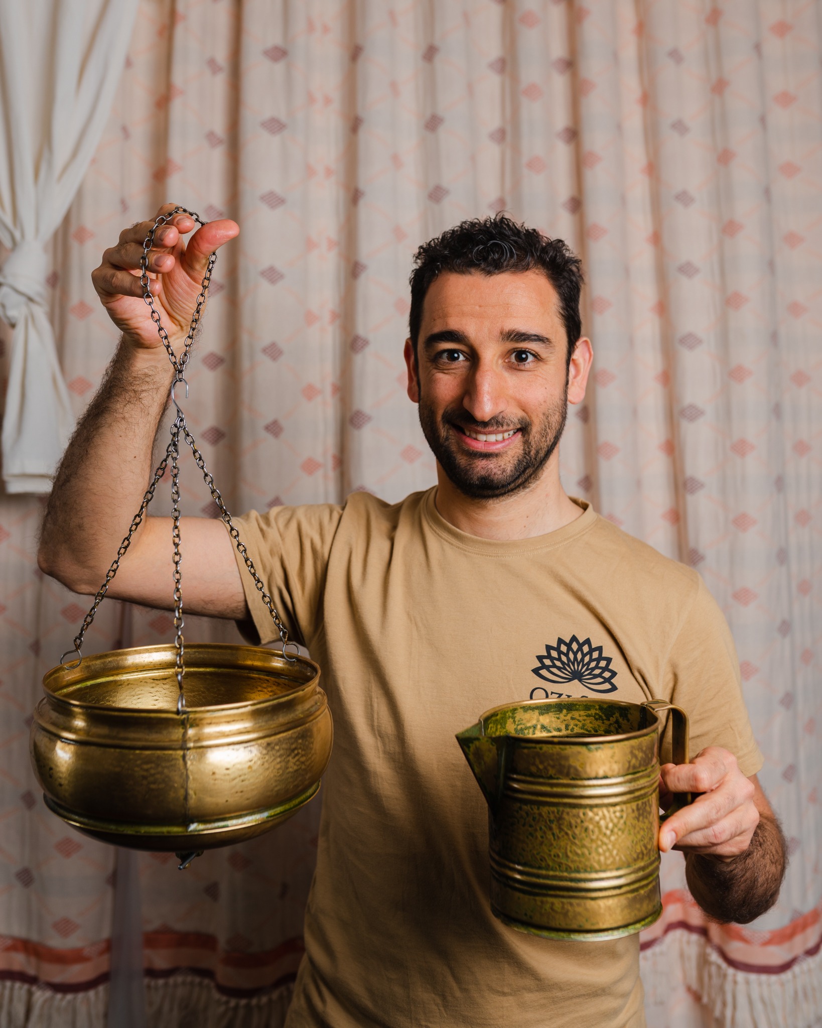Uśmiechnięty mężczyzna w beżowej koszulce z logo trzyma w rękach mosiężne naczynia do ajurwedyjskiego zabiegu, jedno wiszące na łańcuchach, drugie z uchwytem i dzióbkiem.