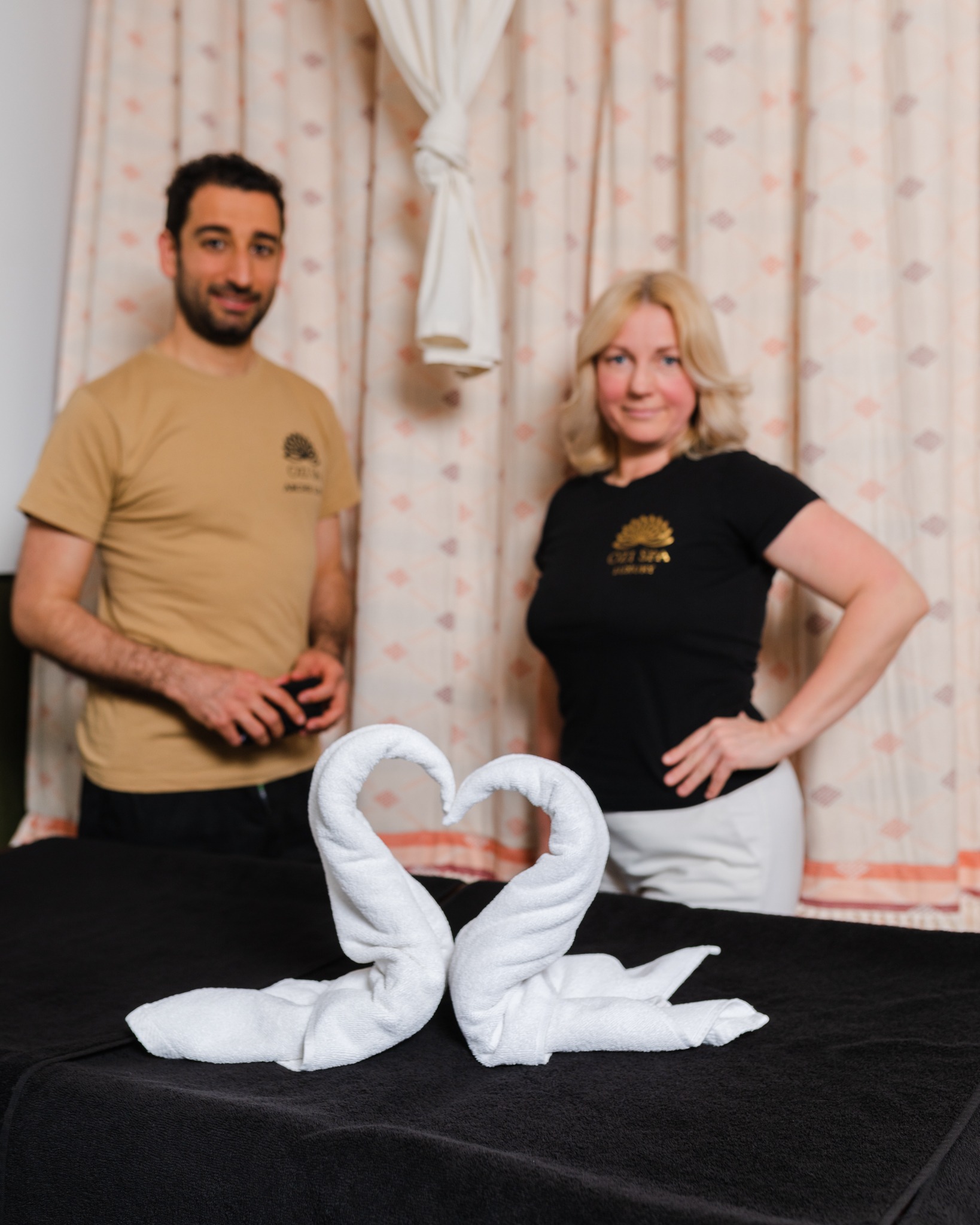 Dwie osoby w koszulkach z logo firmy stoją za łóżkiem do masażu z ręcznikami ułożonymi w kształt łabędzi, tworzących serce.