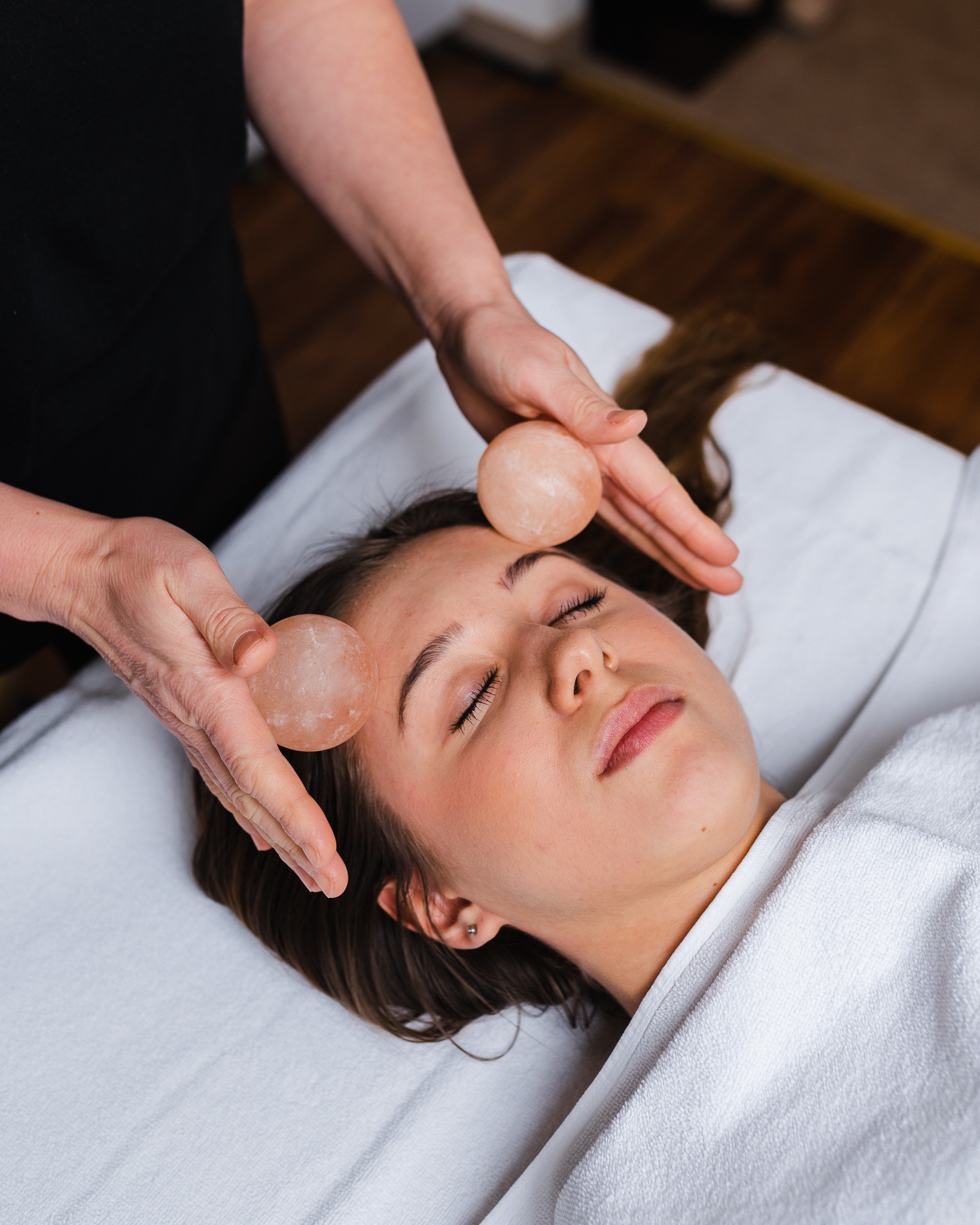 Kobieta leży na białym ręczniku podczas zabiegu na twarz z użyciem ciepłych, różowych kamieni, trzymanych przez dłonie terapeuty w czarnym uniformie.