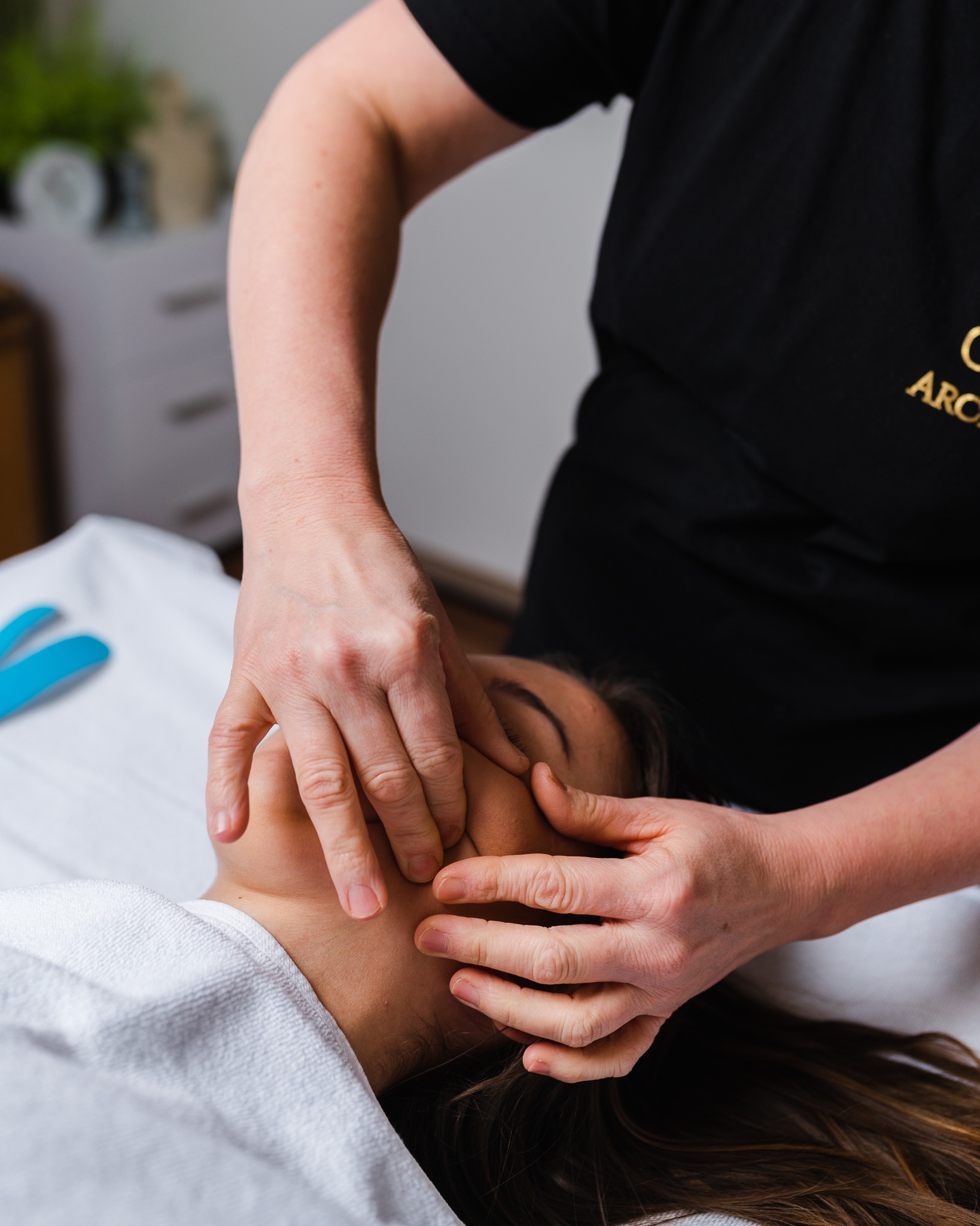 Kobieta podczas masażu twarzy, terapeuta w czarnej koszulce z logo, biały ręcznik frotte, niebieskie aplikatory w tle.