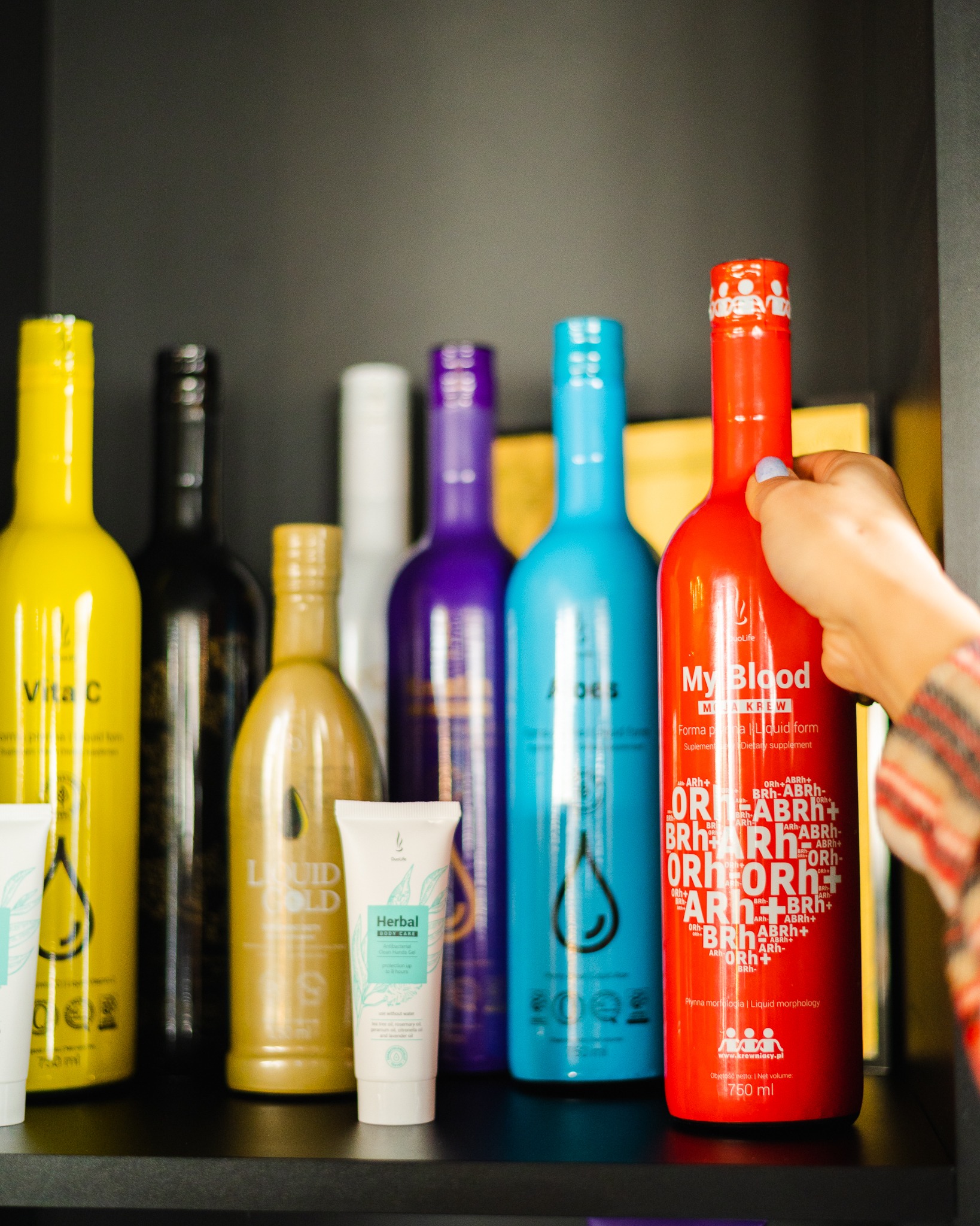 Półka z kolorowymi butelkami i tubkami suplementów diety i kosmetyków, w tym czerwona butelka trzymana przez dłoń, z widocznymi napisami i logo producenta.