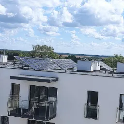 Biały budynek z balkonami i panelami słonecznymi na dachu, widok na odległy krajobraz pod błękitnym niebem z chmurami.