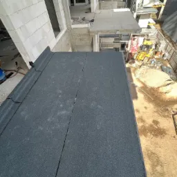 Widok z góry na fragment dachu pokrytego papą termozgrzewalną, z widocznymi łączeniami i fragmentami budynku w surowym stanie oraz placem budowy z piaskiem i sprzętem budowlanym w tle.